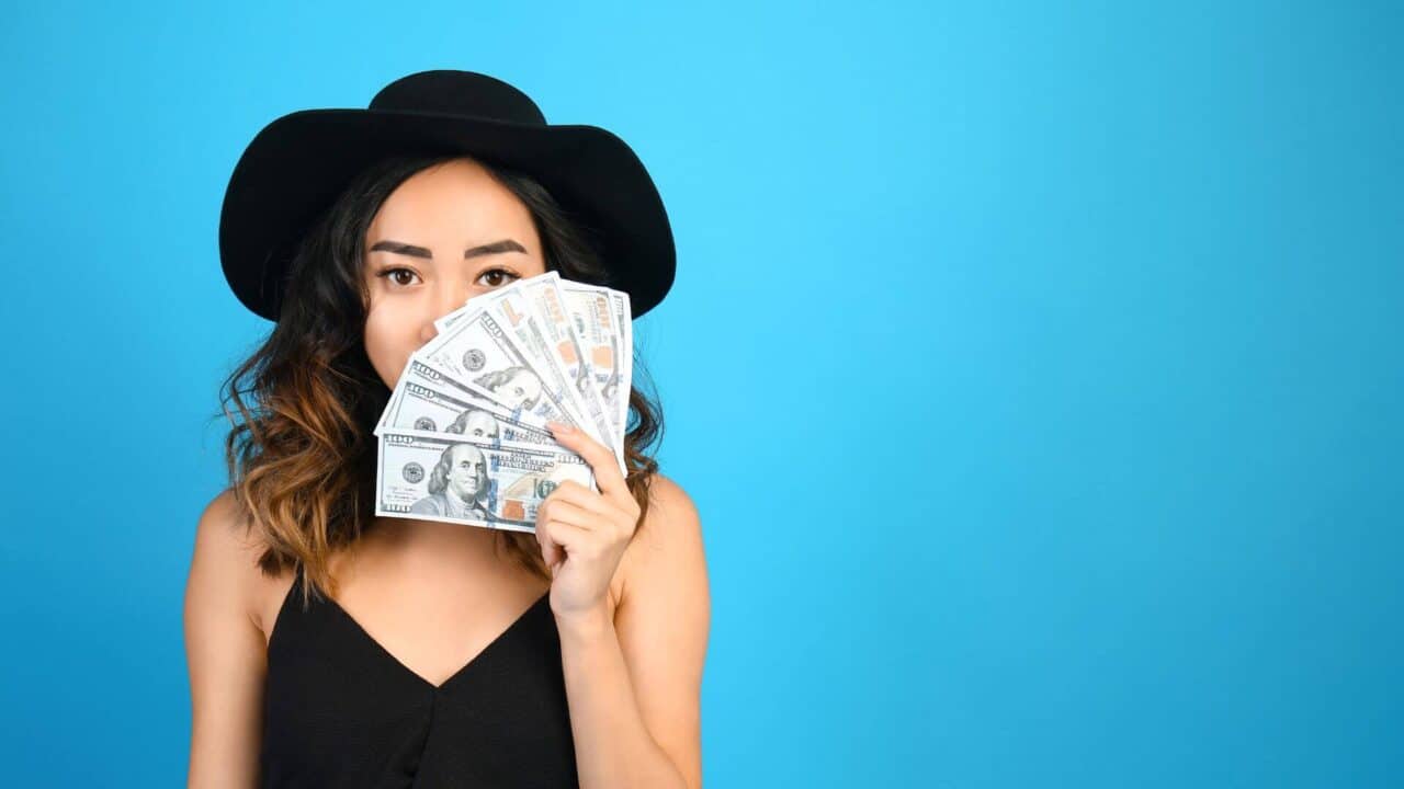 Asian woman wearing a black hat and black tank top holding money blue background