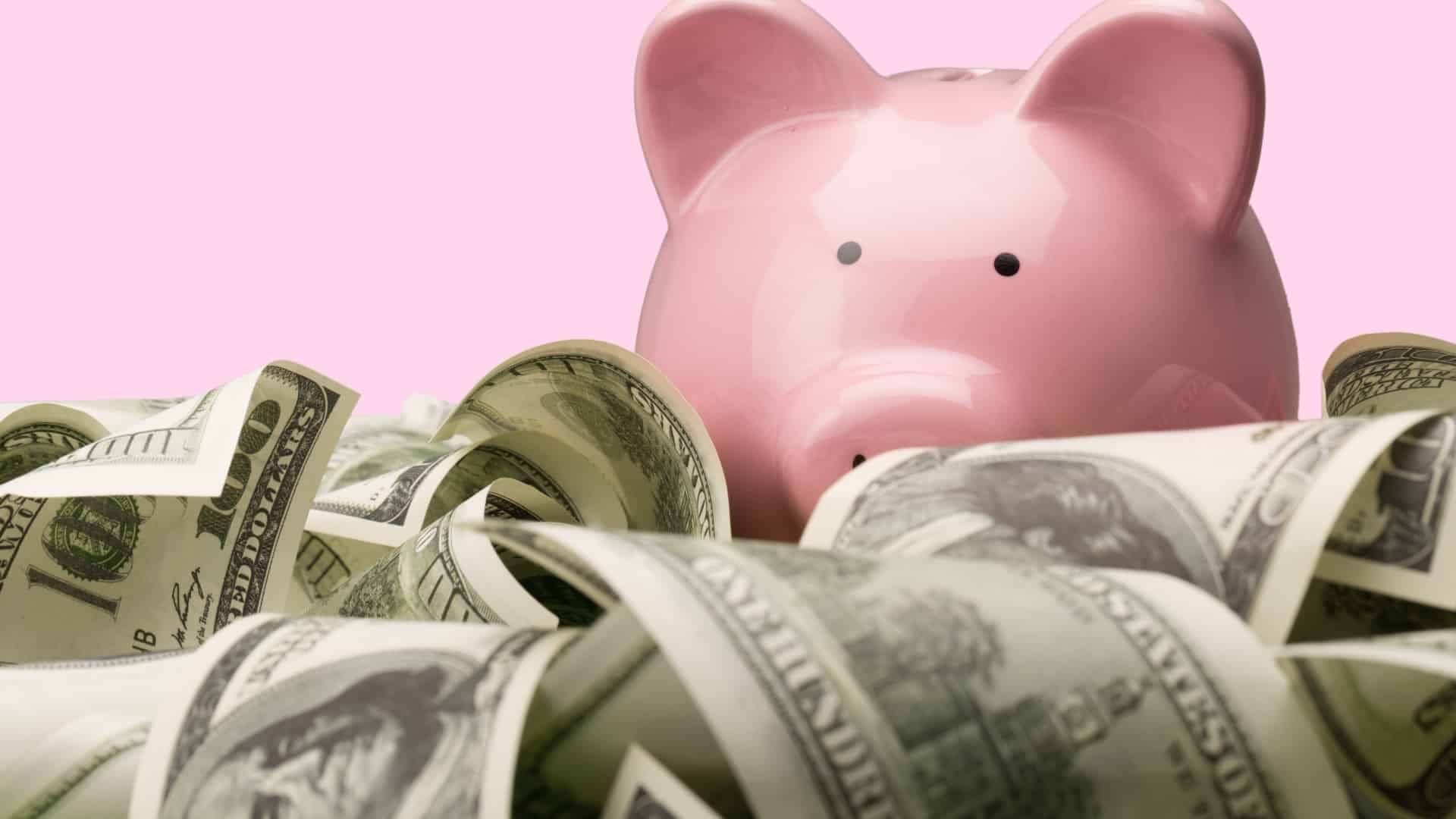 image of pink piggy bank surrounded by a pile on 100 dollar bills pink background
